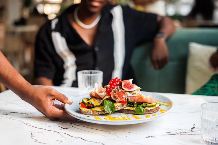 A hand is placing a plate of colorful bruschetta on a marble table in a chic restaurant, with an out-of-focus figure in the background.