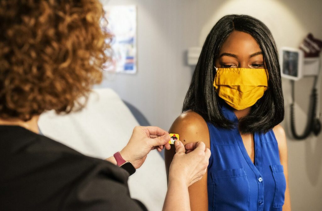 Young Black woman wearing a yellow mask getting a bandaid after a vaccine.