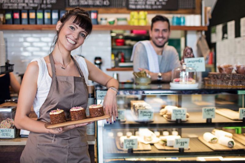 A man and a woman welcome a guest to their coffee shop with a big smile.