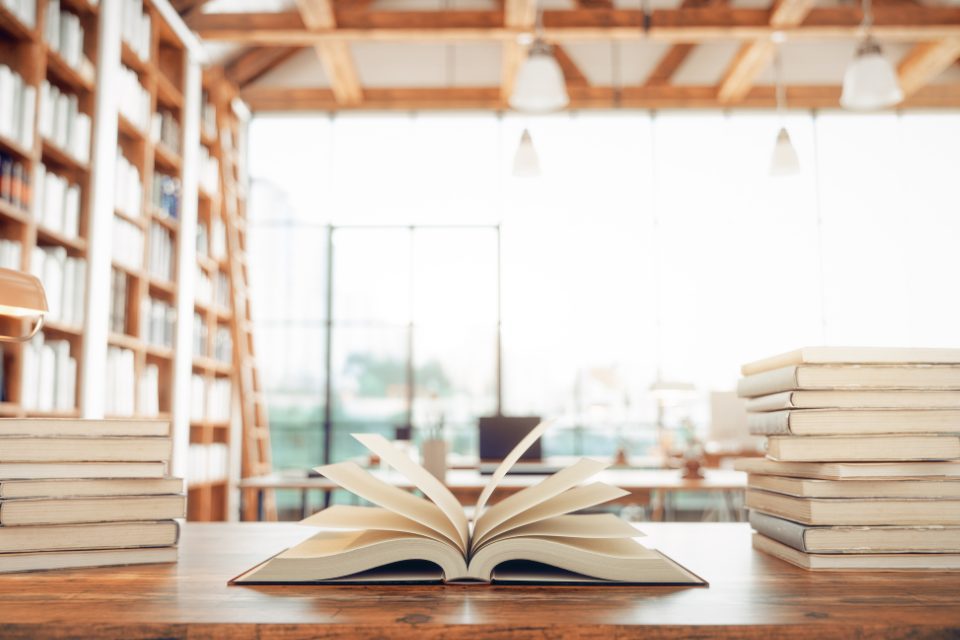 An open book and other books on a table in a sunny library.