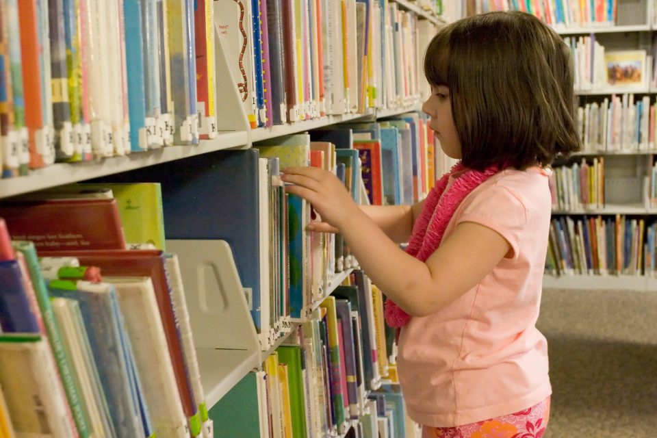 A little girl looking at books on a shelf at a library.