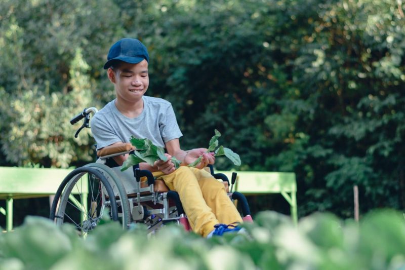 Child in a wheelchair wearing a hat and smiling while checking out a garden in a park.