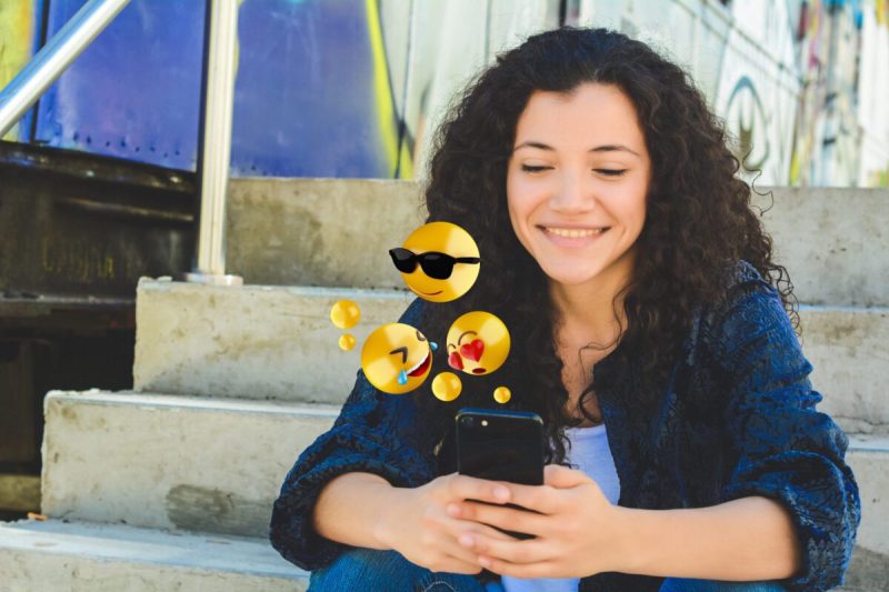 a woman sits smiling at her phone as a few emojis dance in the air